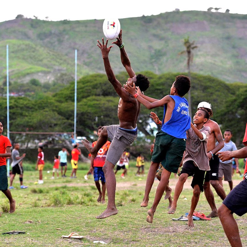 Young players leap for a rugby league ball on a village field below green hills